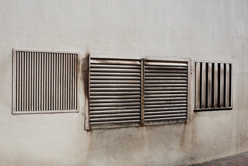 A textured white wall features four rectangular metal ventilation grilles. Two wider grilles in the center have parallel horizontal slats, while the outer two shorter ones have vertical or diagonal slats. The area around some grilles shows signs of dirt and wear, highlighting the need for HVAC cleaning.