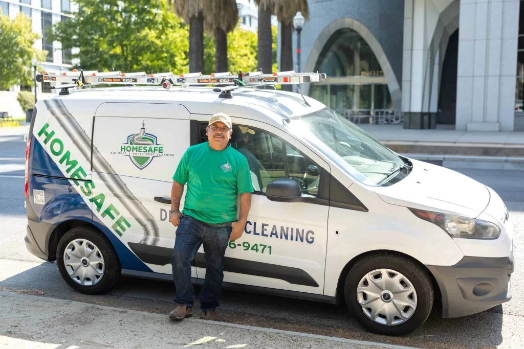 A person in a green "Homesafe" t-shirt and cap stands smiling in front of a "Homesafe" branded van equipped with ladders on its roof. The van is parked on a street with a building and palm trees in the background. Text on the van reads "Dryer Vent Cleaning" and "Exhaust Duct Cleaning.