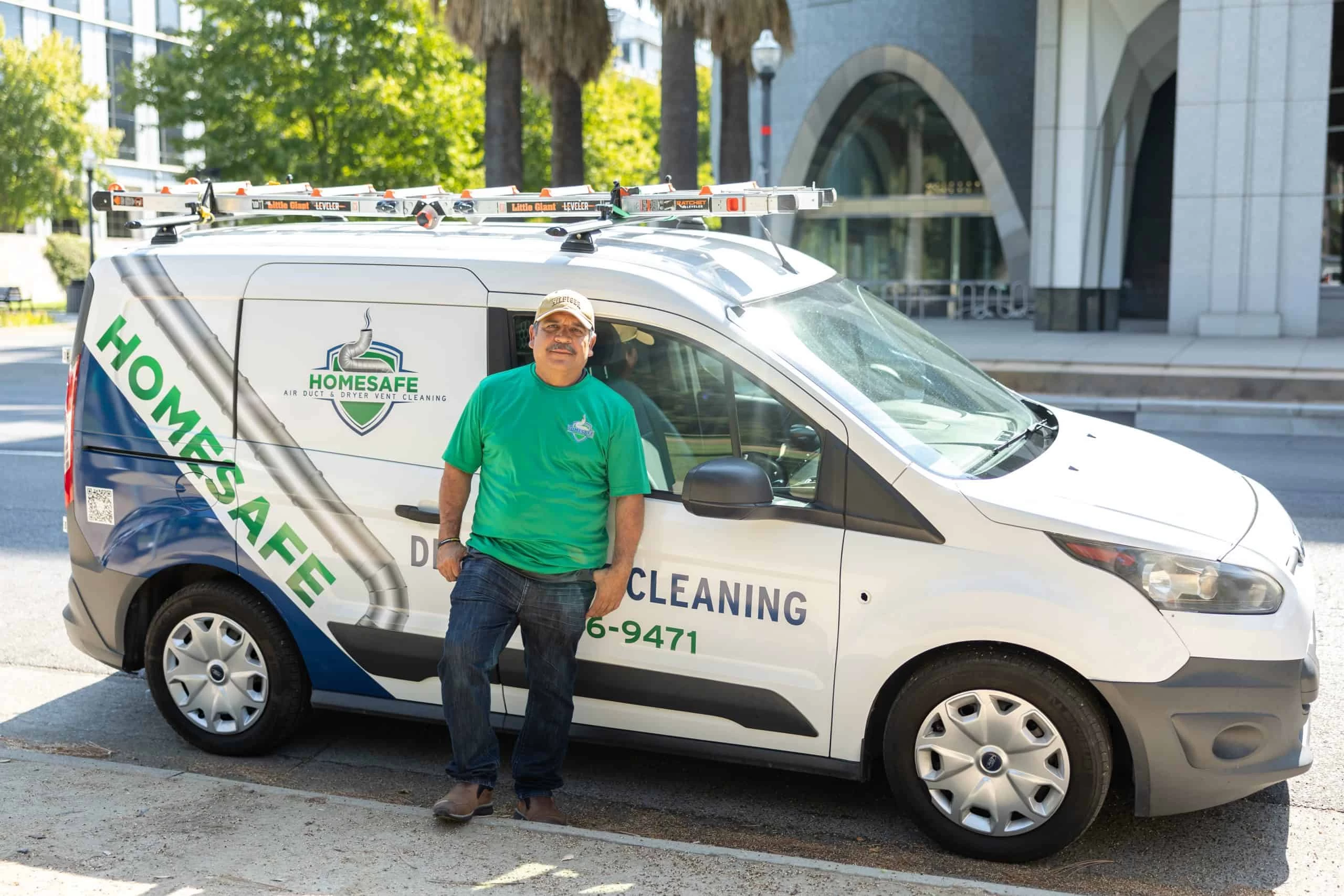 A person in a green "Homesafe" t-shirt and cap stands smiling in front of a "Homesafe" branded van equipped with ladders on its roof. The van is parked on a street with a building and palm trees in the background. Text on the van reads "Dryer Vent Cleaning" and "Exhaust Duct Cleaning.