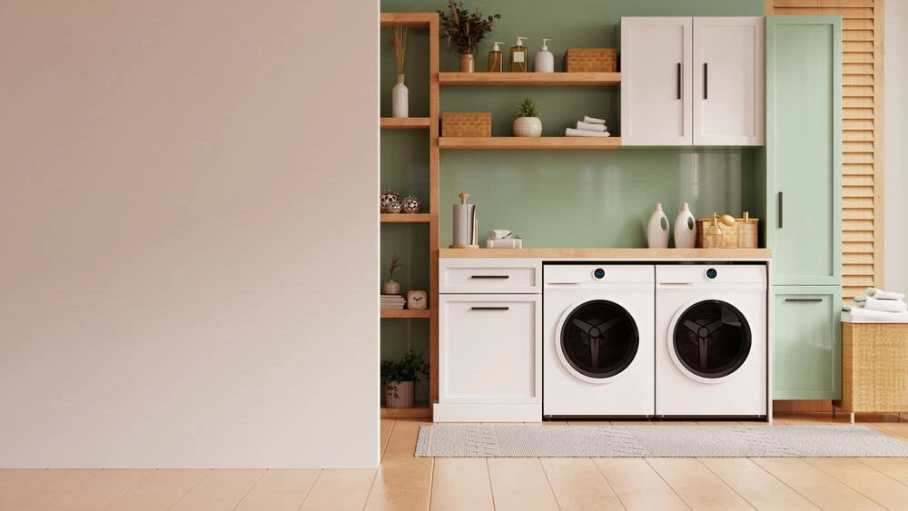 A modern laundry room features a washer and dryer with cabinet storage above and to the side. Wooden shelves hold decorative items, and a green accent wall adds color. A woven basket and towels are placed near the window with wooden blinds, emphasizing the importance of regular dryer vent cleaning.