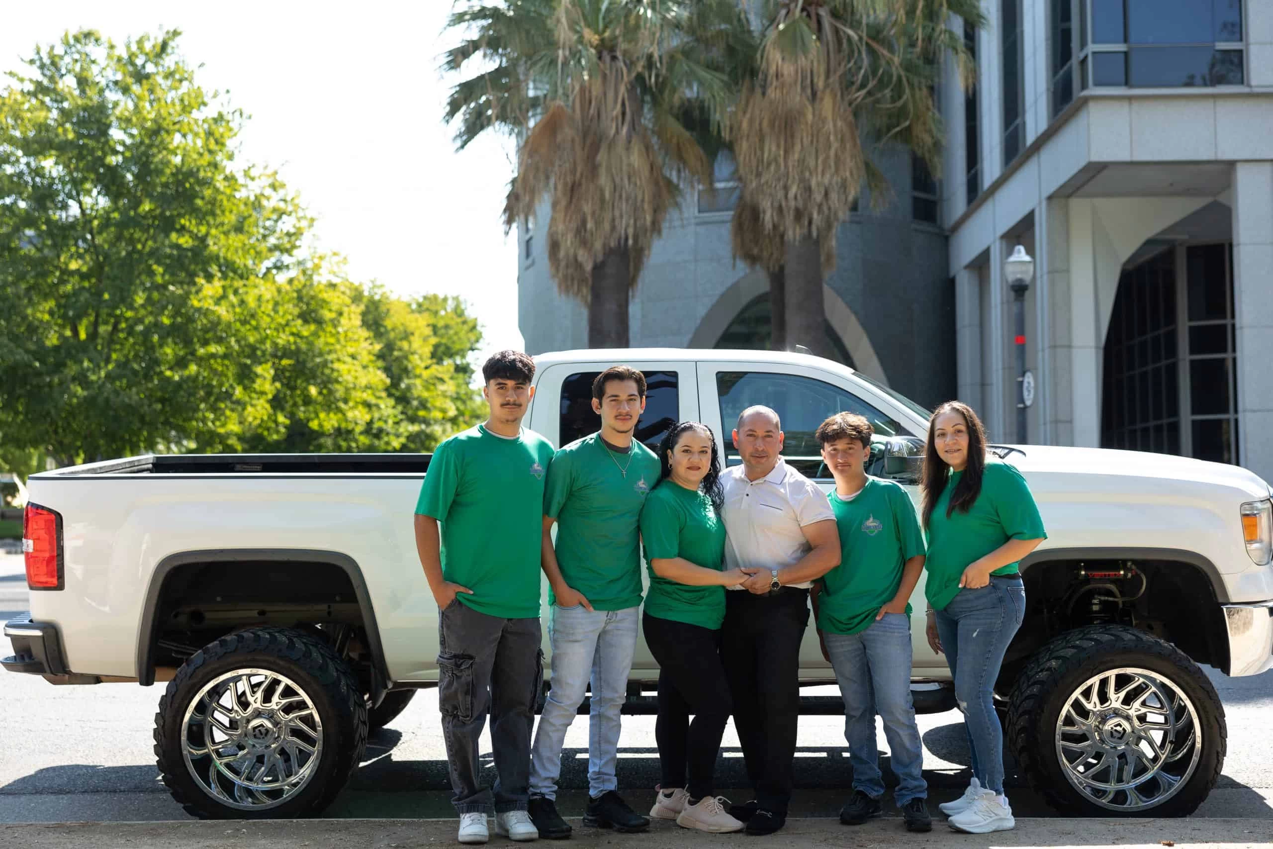A group of six people wearing green shirts stand in front of a white pickup truck with large chrome wheels. Behind them are palm trees and a building, possibly indicating a successful HVAC cleaning job. Four young men and two women pose together, some with arms around each other. It's a sunny day.