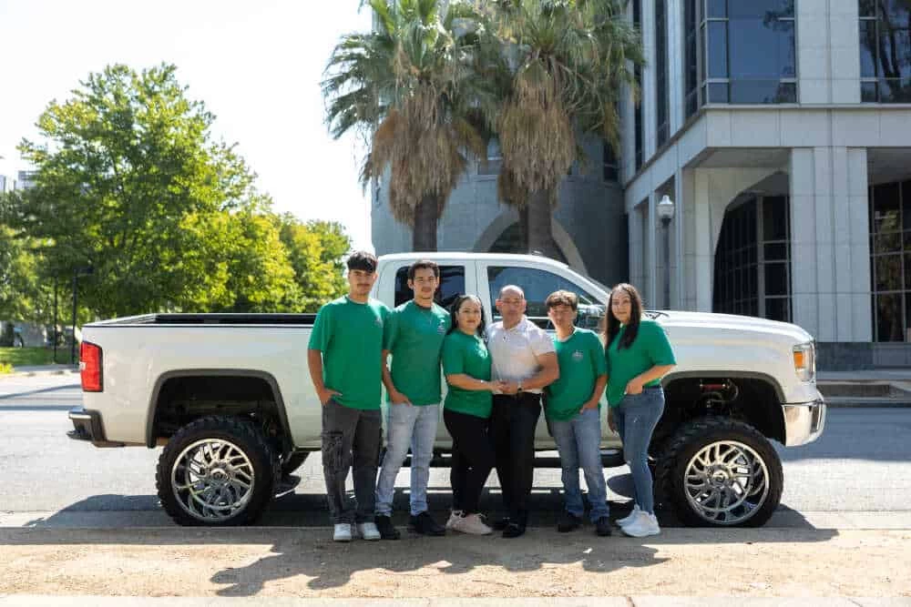 A group of six people wearing green shirts pose in front of a large, white pickup truck. They are standing on a street with trees and a modern building, featuring large windows and palm trees in the background—ready for their next HVAC system maintenance job.