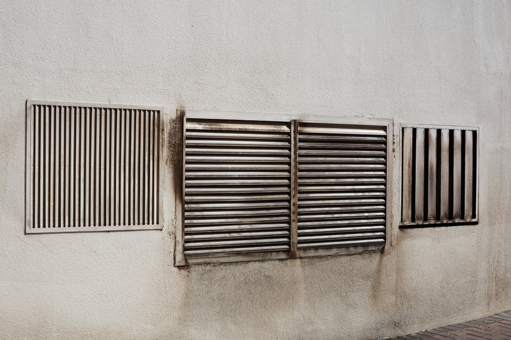 A textured white wall features four rectangular metal ventilation grilles. Two wider grilles in the center have parallel horizontal slats, while the outer two shorter ones have vertical or diagonal slats. The area around some grilles shows signs of dirt and wear, highlighting the need for HVAC cleaning.