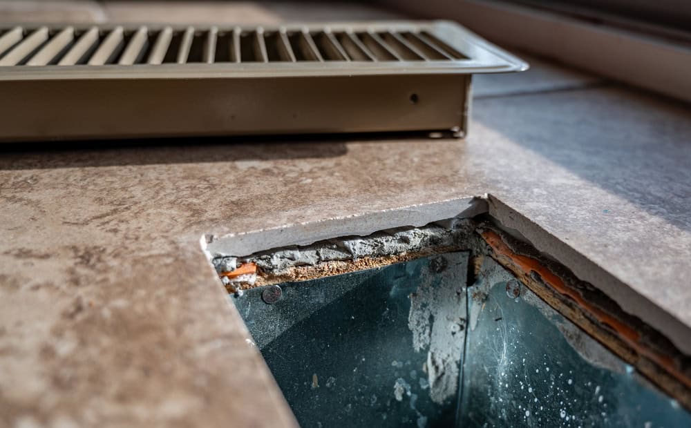 A close-up of a floor vent with the grille removed, revealing the metal ductwork below. The surrounding tile floor shows signs of wear, with chipped edges around the vent opening. The vent cover rests beside the opening, highlighting the need for ventilation system cleaning.