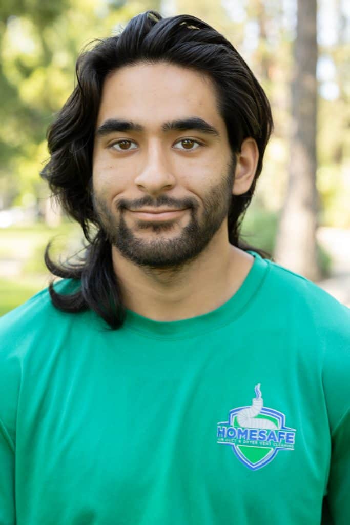 A person with long dark hair and a beard is smiling slightly, capturing the welcoming spirit of our "About Us" ethos. Wearing a green "HOMESAFE" t-shirt, they stand against a blurred background with sunlight filtering through trees.