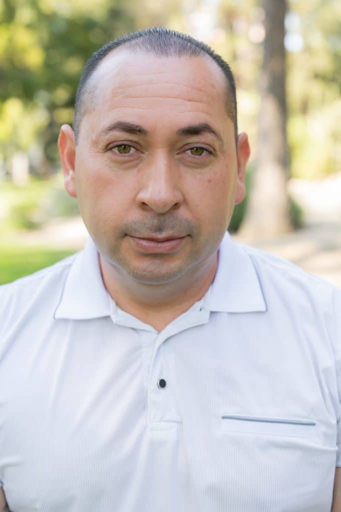 A man with short hair and a white polo shirt stands outdoors, looking directly at the camera for the 'About Us' page. Trees and greenery are blurred in the background, suggesting a park or garden setting.