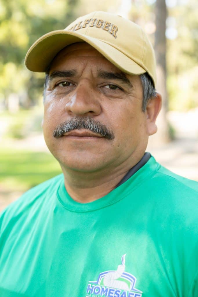 A man in a yellow cap and green shirt stands outdoors, looking directly at the camera. The park setting, with its blurred trees and grass, subtly hints at the essence of our "About Us" narrative—grounded in nature and authenticity.