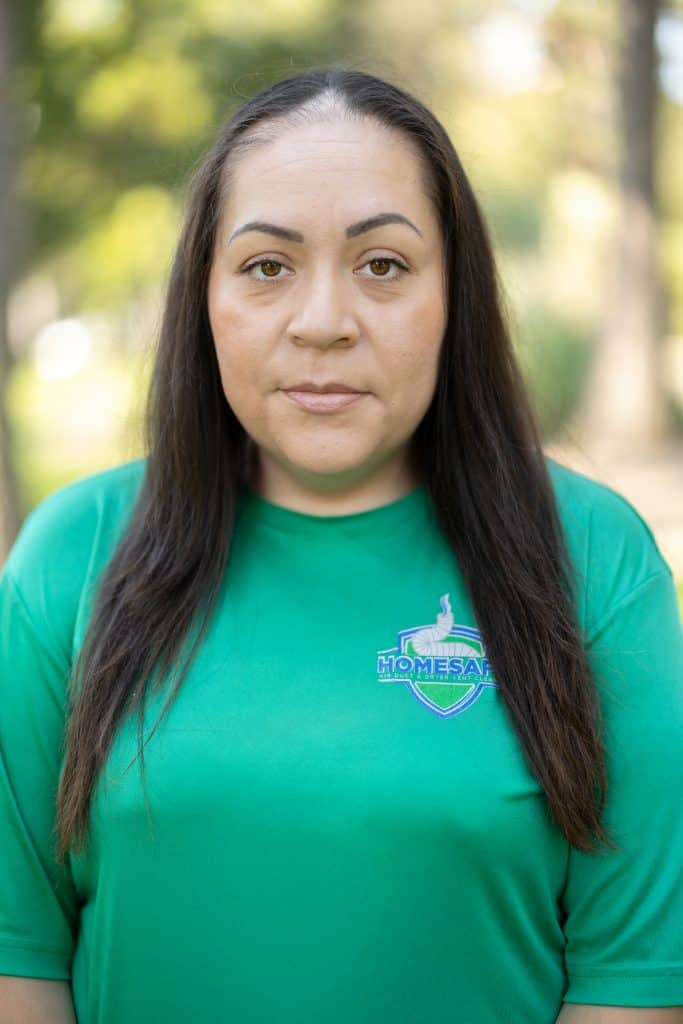 A person with long dark hair stands outdoors, wearing a green shirt with a logo. The blurred background, filled with trees and greenery, hints at a park setting, creating an inviting atmosphere akin to taking a refreshing stroll after an air duct cleaning in Sacramento, CA.
