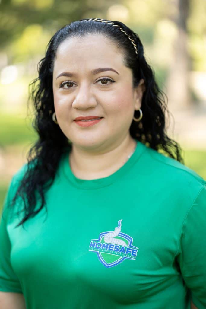 A woman with curly black hair and hoop earrings smiles slightly while standing outdoors, embodying the spirit of "Home Safe". Her green shirt proudly announces “About Us” amidst a backdrop of blurred greenery.