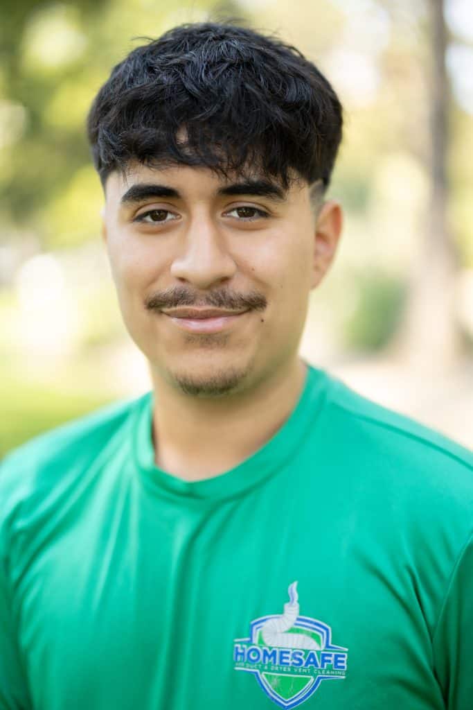 A person in a green "Home Safe" shirt stands outdoors, smiling as if they are the embodiment of our About Us mission. The background is blurred, with hints of greenery and sunlight adding warmth to the scene.