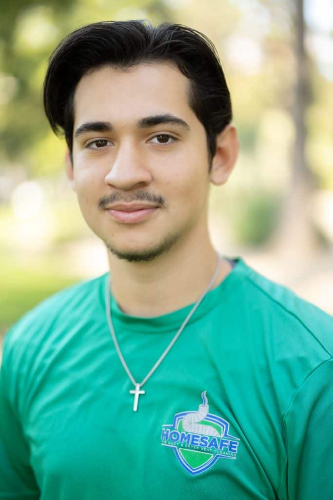 A person with short dark hair and a mustache, wearing a green "HOMESAFE" shirt and a cross necklace, stands outdoors with blurred trees in the background, symbolizing the dedication we embody in our About Us story.