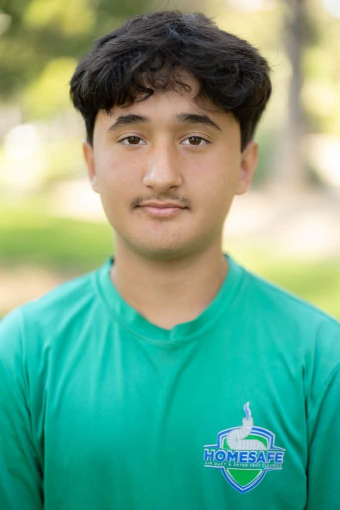 Standing outside amidst blurred greenery, a young person with short, dark hair is confidently sporting a green "HOMESAFE" shirt—a testament to their commitment to our cause. It's as if they're the embodiment of an "About Us" page, sharing our essence at a glance.