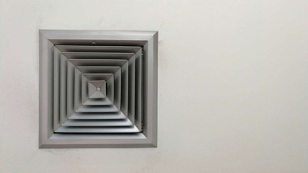 A square metallic air vent is mounted on a plain white wall. The vent has multiple concentric layered frames, creating a geometric pattern. The wall is smooth with some slight shadows around the vent.