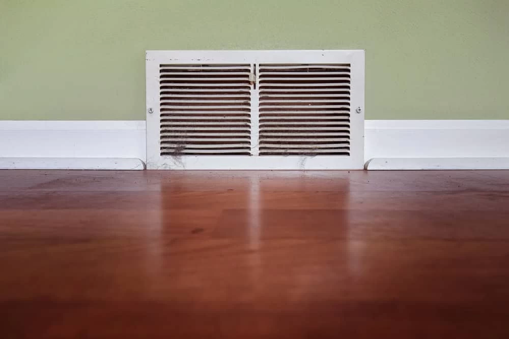 A white air vent with horizontal slats is set in a pale green wall above a wooden floor. Dust is visible on the vent, contrasting with the clean floor and baseboard.
