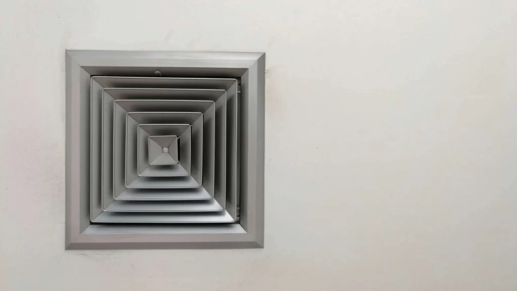 A square metallic air vent is mounted on a plain white wall. The vent has multiple concentric layered frames, creating a geometric pattern. The wall is smooth with some slight shadows around the vent.