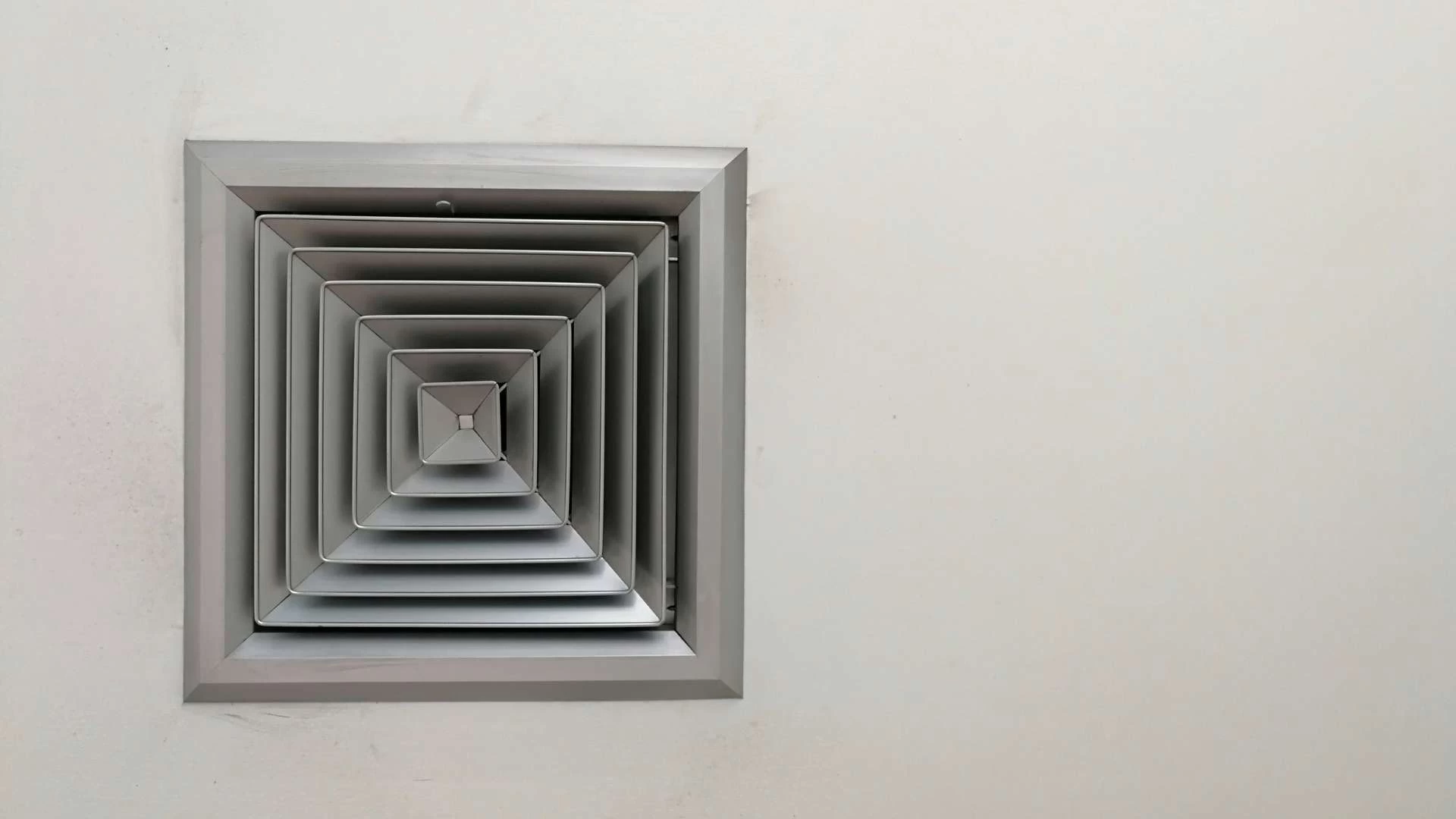 A square metallic air vent is mounted on a plain white wall. The vent has multiple concentric layered frames, creating a geometric pattern. The wall is smooth with some slight shadows around the vent.