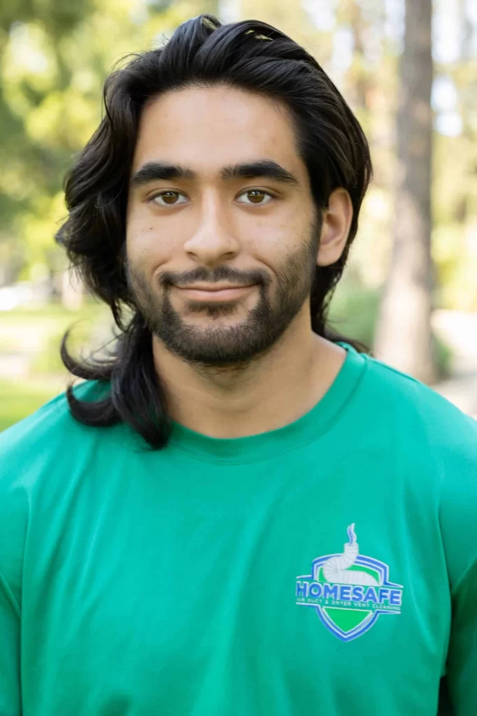 A person with long dark hair and a beard is smiling slightly, capturing the welcoming spirit of our "About Us" ethos. Wearing a green "HOMESAFE" t-shirt, they stand against a blurred background with sunlight filtering through trees.