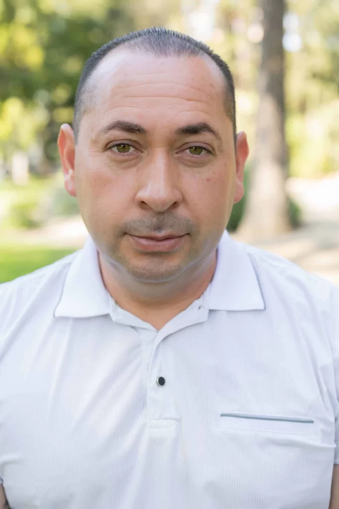 A man with short hair and a white polo shirt stands outdoors, looking directly at the camera for the 'About Us' page. Trees and greenery are blurred in the background, suggesting a park or garden setting.