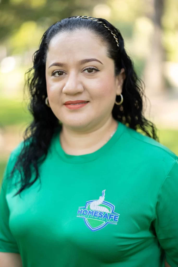 A woman with curly black hair and hoop earrings smiles slightly while standing outdoors, embodying the spirit of "Home Safe". Her green shirt proudly announces “About Us” amidst a backdrop of blurred greenery.