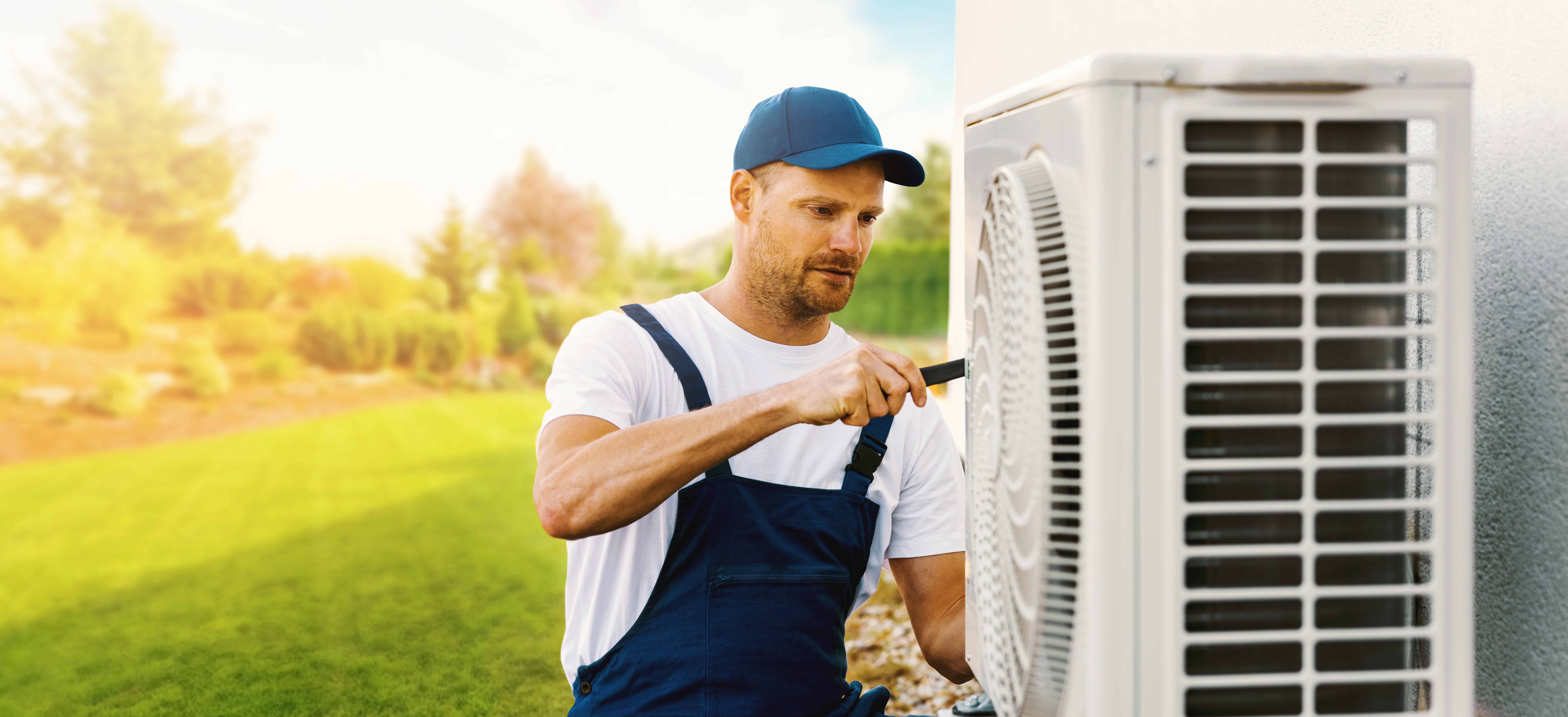 A man in a blue cap and overalls is servicing an outdoor air conditioning unit with a screwdriver. The scene is set in a sunny garden with a green lawn and blurred trees in the background.