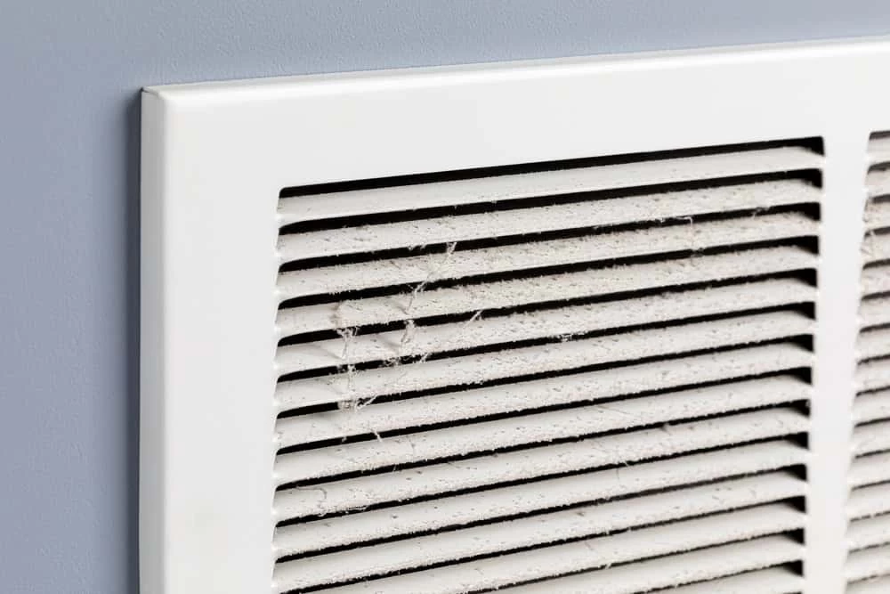 A close-up of a dusty air vent on a gray wall, with dust and cobwebs clinging to the slats of the white vent cover.