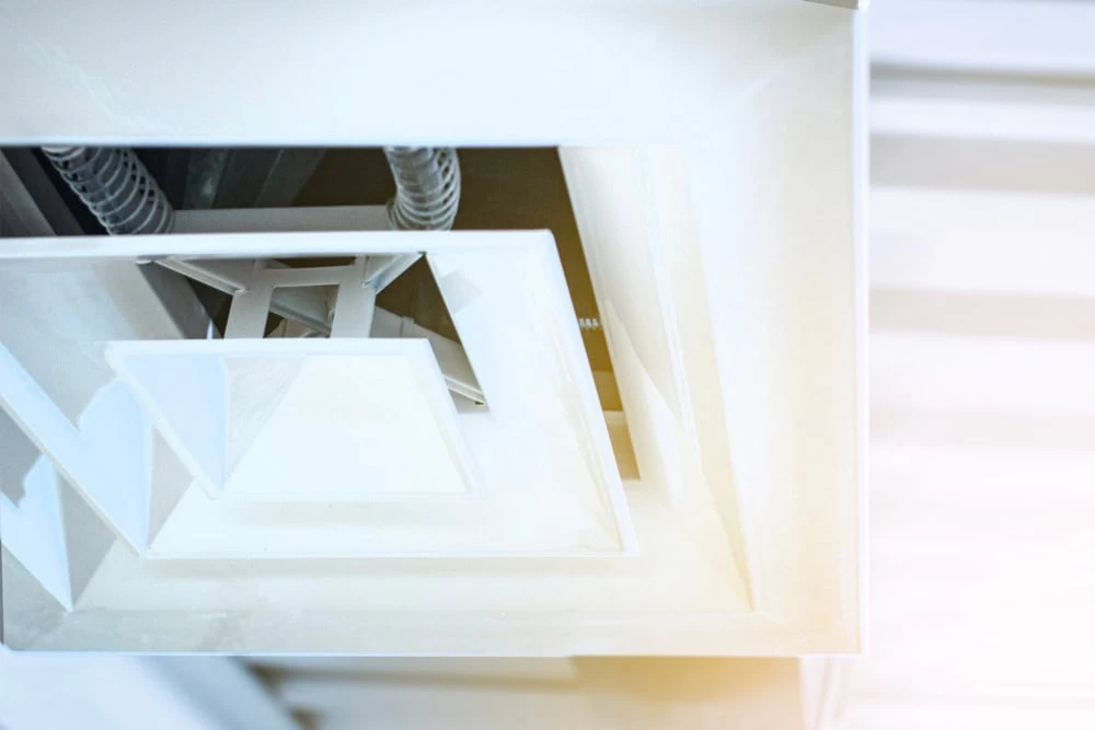 Close-up of a white ceiling air vent with geometric patterns. The vent has several overlapping square layers with visible ductwork in the background. Bright light filters through, giving a clean and modern appearance.