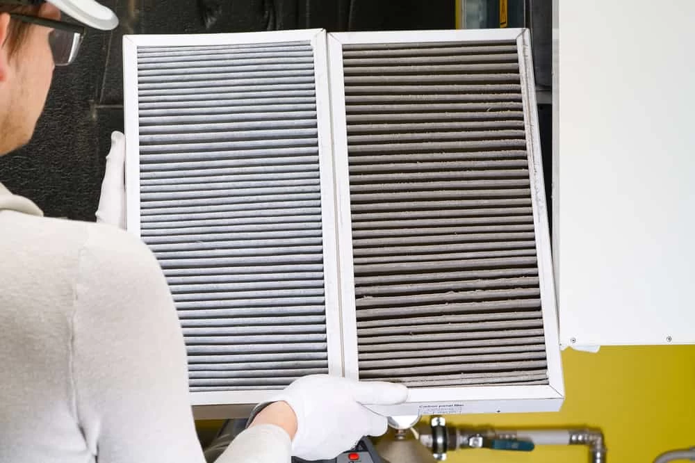 A person wearing gloves holds two air filters. The left filter is clean and grey, while the right one is dirty and dark with dust. The background includes a wall and a small section of industrial equipment.