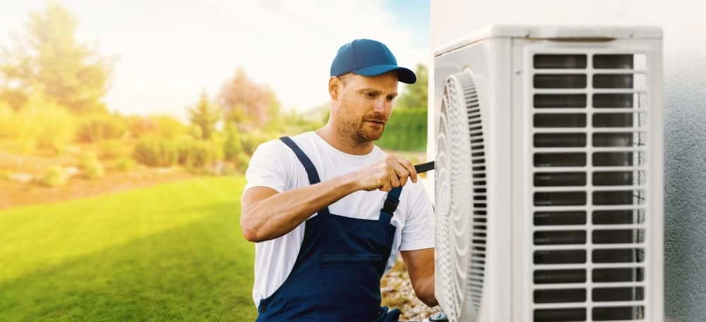 A man in a blue cap and overalls is servicing an outdoor air conditioning unit with a screwdriver. The scene is set in a sunny garden with a green lawn and blurred trees in the background.