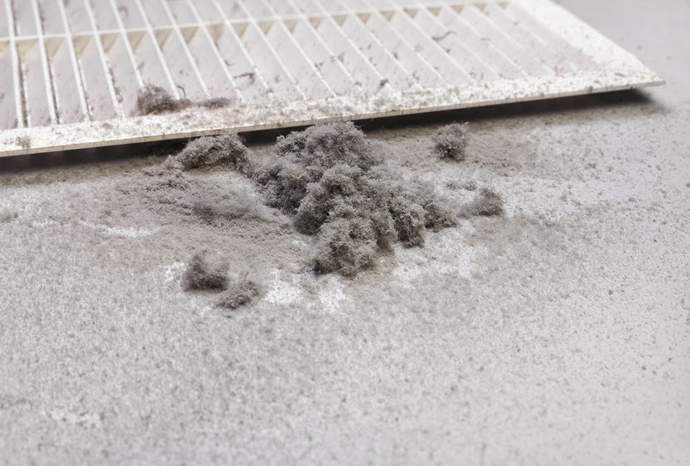 A close-up of a dirty air filter with accumulated dust and debris. The filter is lying flat on a surface, and the dust is visibly clumped on and around it, highlighting the need for cleaning or replacement.