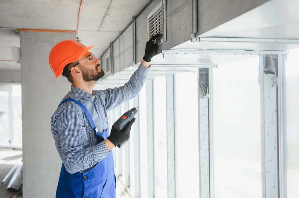 A construction worker wearing an orange hard hat, safety glasses, and blue overalls is using a drill to install or repair a vent on a large air duct system inside a building under construction or renovation.