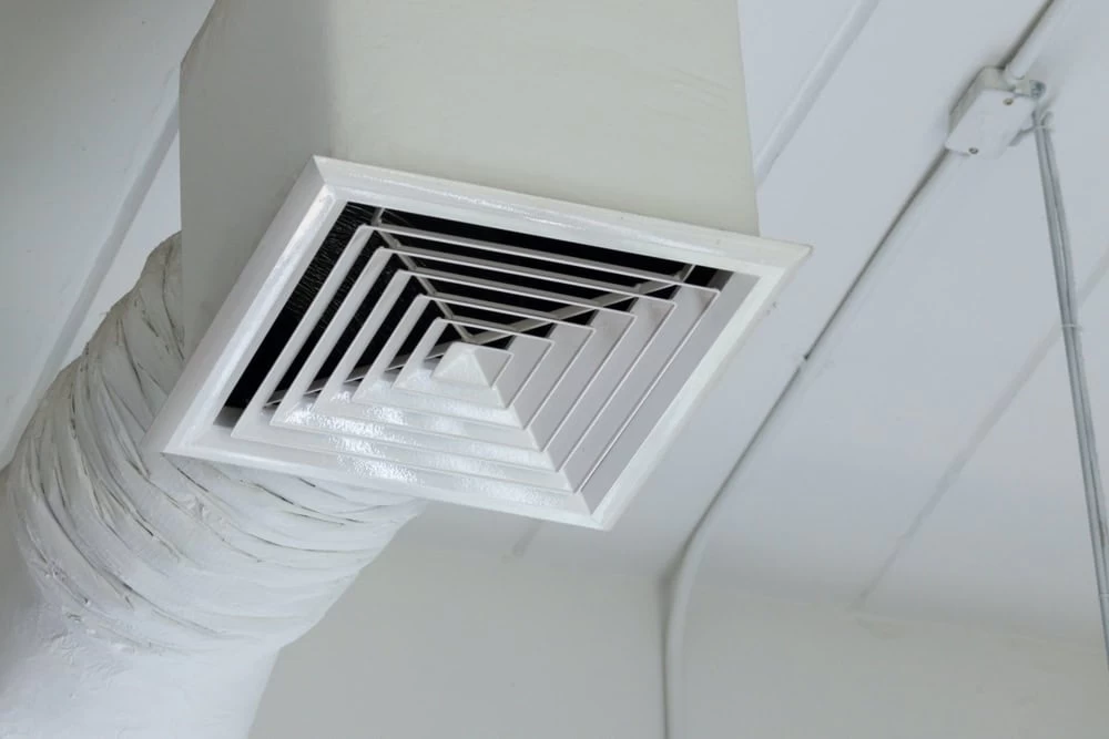 Close-up view of a white, square ceiling air vent with a spiral design. The vent is part of an exposed duct system, set against a white ceiling background, and is slightly angled.