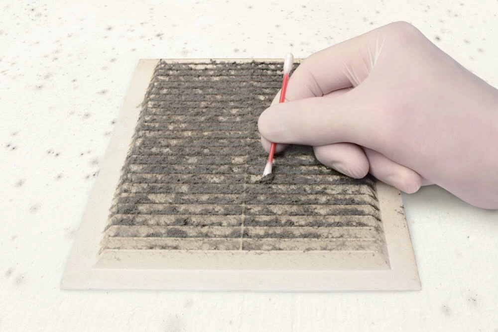 A gloved hand uses a cotton swab to clean a dirty, dusty air vent. The surface is covered with black particles.
