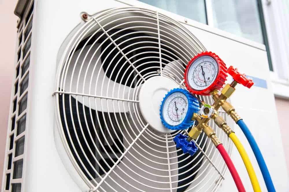 Close-up of an air conditioning unit with pressure gauges attached. The gauges have blue and red dials and are connected by colorful hoses. The fan of the unit is visible, and there is a building wall in the background.
