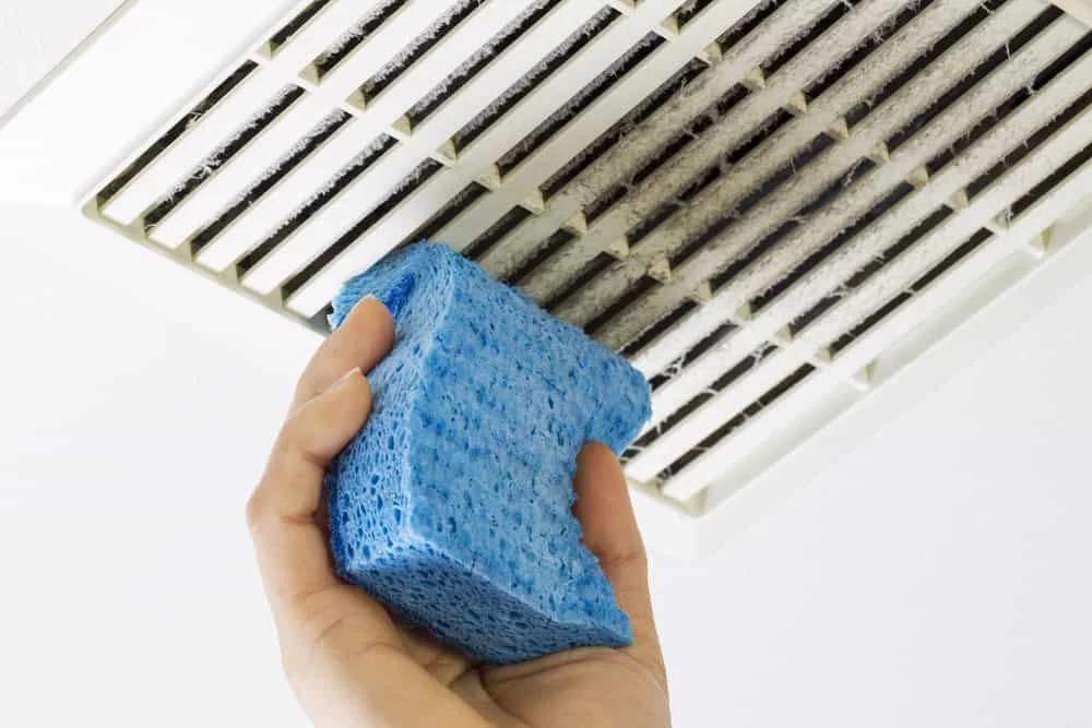 A hand holding a blue sponge diligently cleans a dusty ceiling air vent, highlighting the importance of regular maintenance like air duct cleaning in Sacramento, CA.