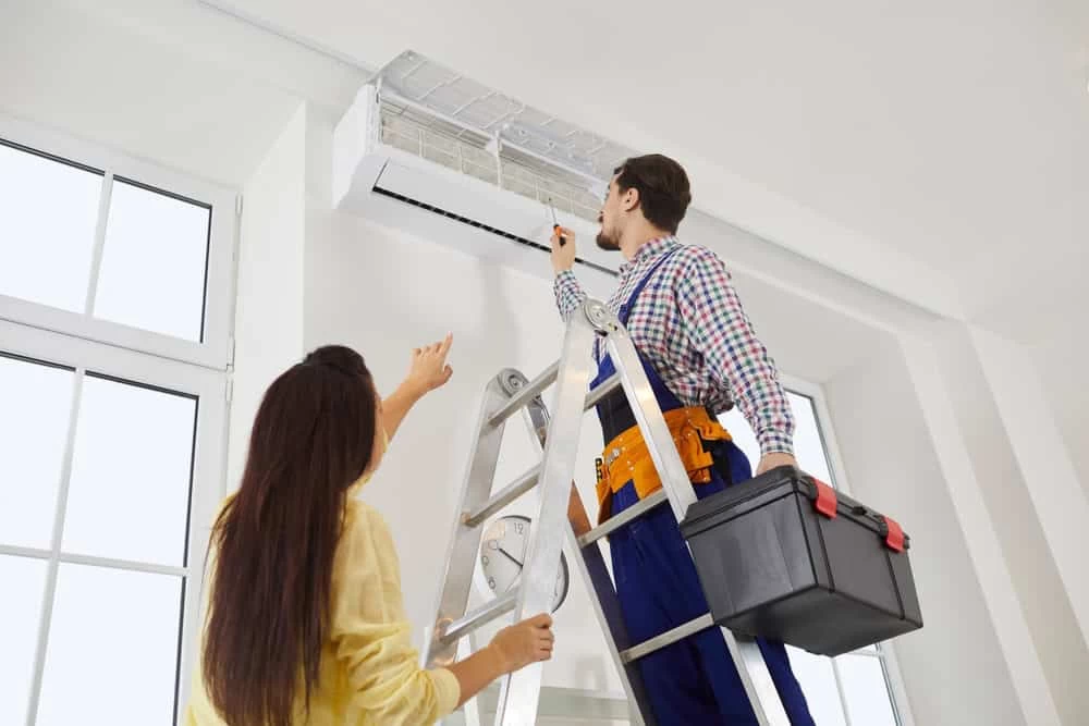 A technician on a ladder repairs an air conditioner while holding a toolbox. A woman stands nearby, pointing and discussing the work. They are in a bright room with large windows.