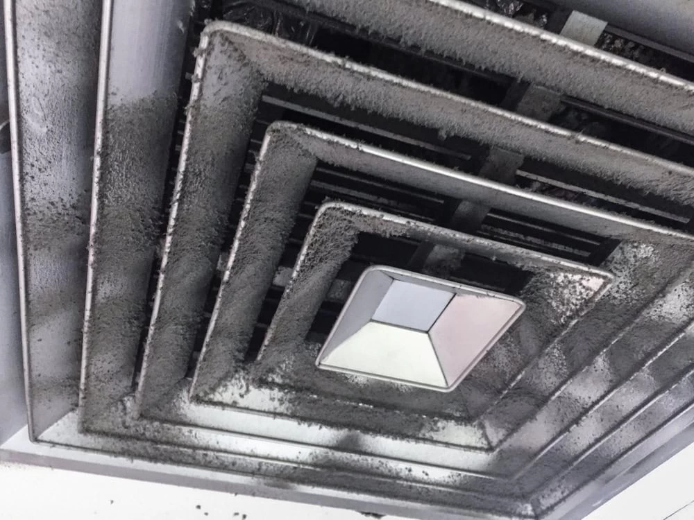 Close-up of a ceiling air vent covered in dust and dirt. The vent is square-shaped with multiple layers. Dust has accumulated heavily on the edges and surfaces, indicating it hasn't been cleaned in a while.