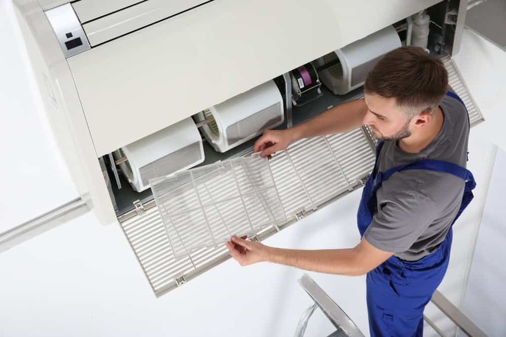 A person in a gray shirt and blue overalls stands on a ladder, cleaning or inspecting a filter from an air conditioning unit mounted on a wall.