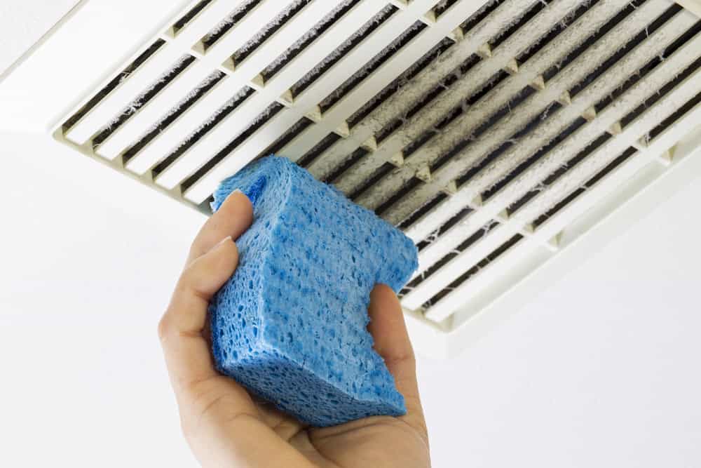A hand holding a blue sponge diligently cleans a dusty ceiling air vent, highlighting the importance of regular maintenance like air duct cleaning in Sacramento, CA.
