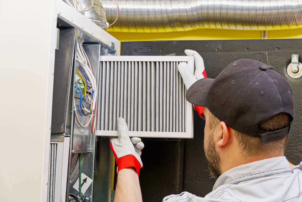 A technician wearing gloves and a cap installs an air filter into an HVAC system. The background shows ductwork and various wires inside the unit.