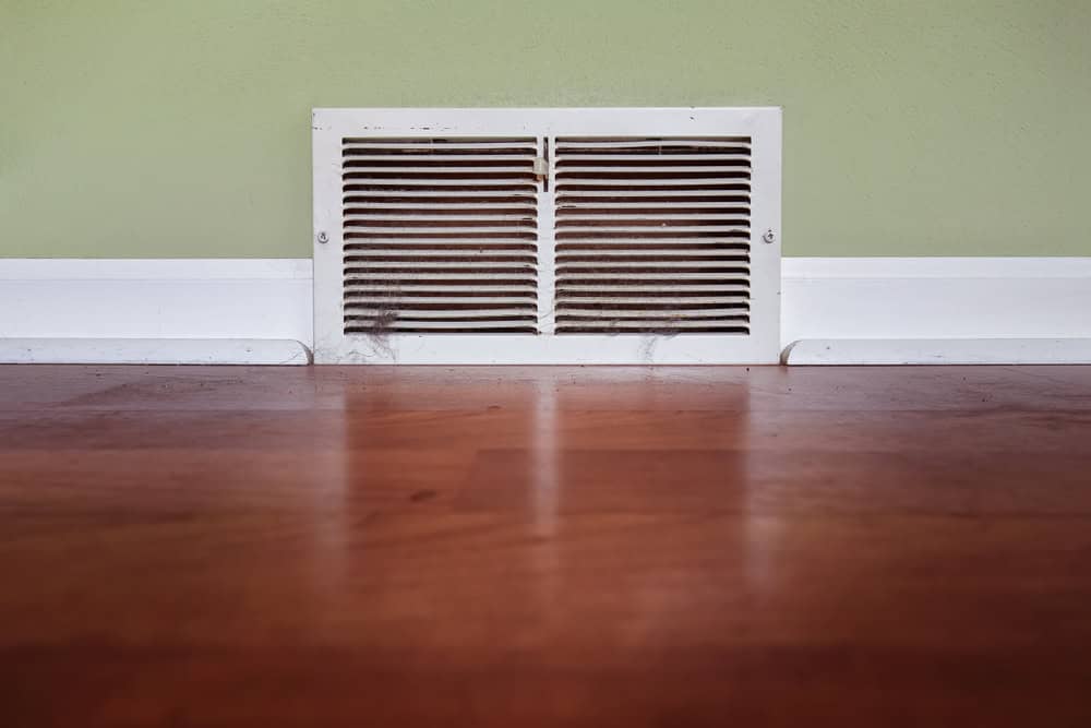 A white air vent with horizontal slats is set in a pale green wall above a wooden floor. Dust is visible on the vent, contrasting with the clean floor and baseboard.