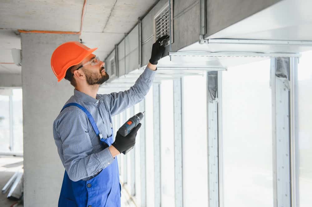A construction worker wearing an orange hard hat, safety glasses, and blue overalls is using a drill to install or repair a vent on a large air duct system inside a building under construction or renovation.