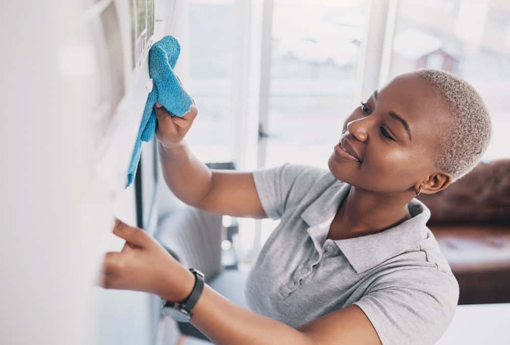 A person wearing a gray polo shirt is smiling while cleaning a whiteboard with a blue cloth. They have short, light-colored hair and are standing in a bright room with large windows and a sofa in the background.