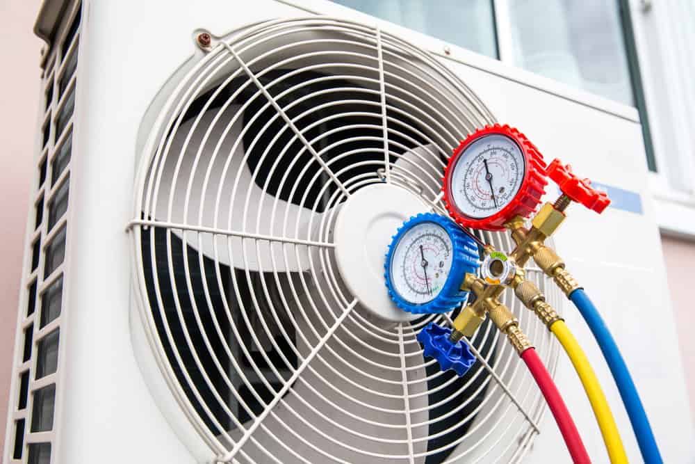 Close-up of an air conditioning unit with pressure gauges attached. The gauges have blue and red dials and are connected by colorful hoses. The fan of the unit is visible, and there is a building wall in the background.