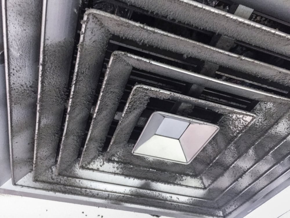 Close-up of a ceiling air vent covered in dust and dirt. The vent is square-shaped with multiple layers. Dust has accumulated heavily on the edges and surfaces, indicating it hasn't been cleaned in a while.
