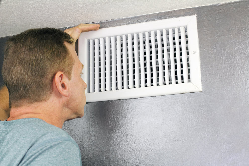 A person inspects a white air vent on a gray wall. They are examining it closely, possibly checking for airflow or maintenance issues.