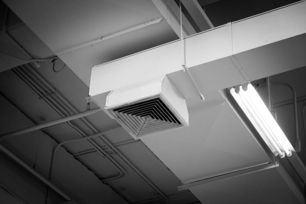 Black and white image of an industrial ceiling featuring an air vent and fluorescent light fixture. The ceiling shows exposed pipes and structural elements.