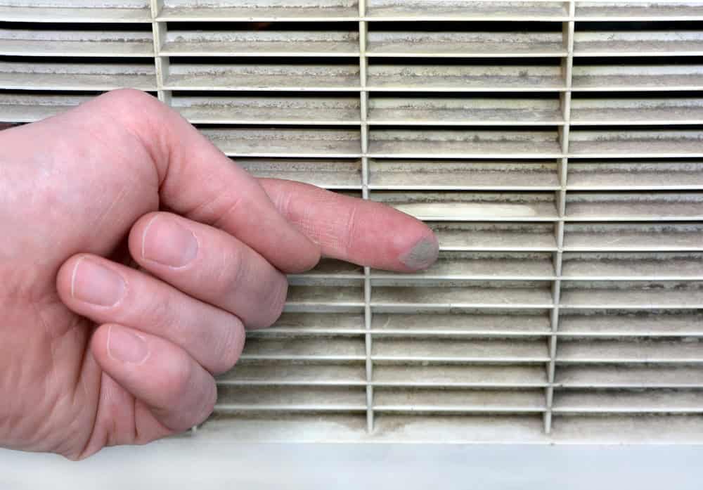 A finger touches a dusty air vent, showing the accumulated dirt on the fingertip. The vent features horizontal slats and the background emphasizes the need for cleaning.