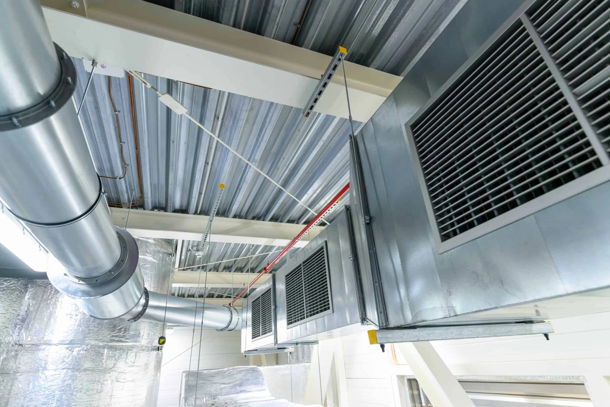 Industrial ceiling with large metal ventilation ducts and air vents. The ducts are connected to a corrugated metal roof, and some parts are insulated with shiny metallic material. The scene appears to be within a commercial or warehouse space.