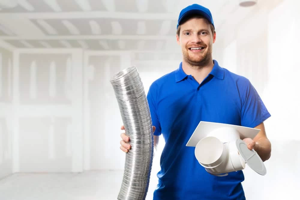 A man in a blue shirt and cap smiles while holding a large coil of ductwork in his right hand and ventilation parts in his left. He stands in an unfinished room with drywall.