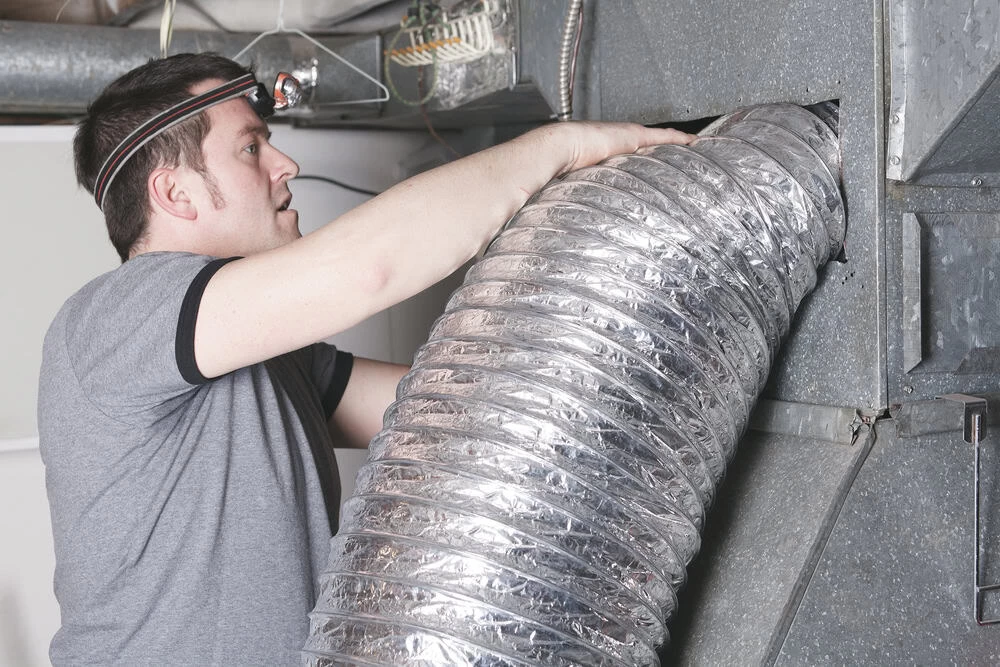 A person wearing a headlamp is inspecting or repairing a large silver flexible duct connected to a ventilation system. They are indoors, and the setting appears to be industrial or related to HVAC maintenance.